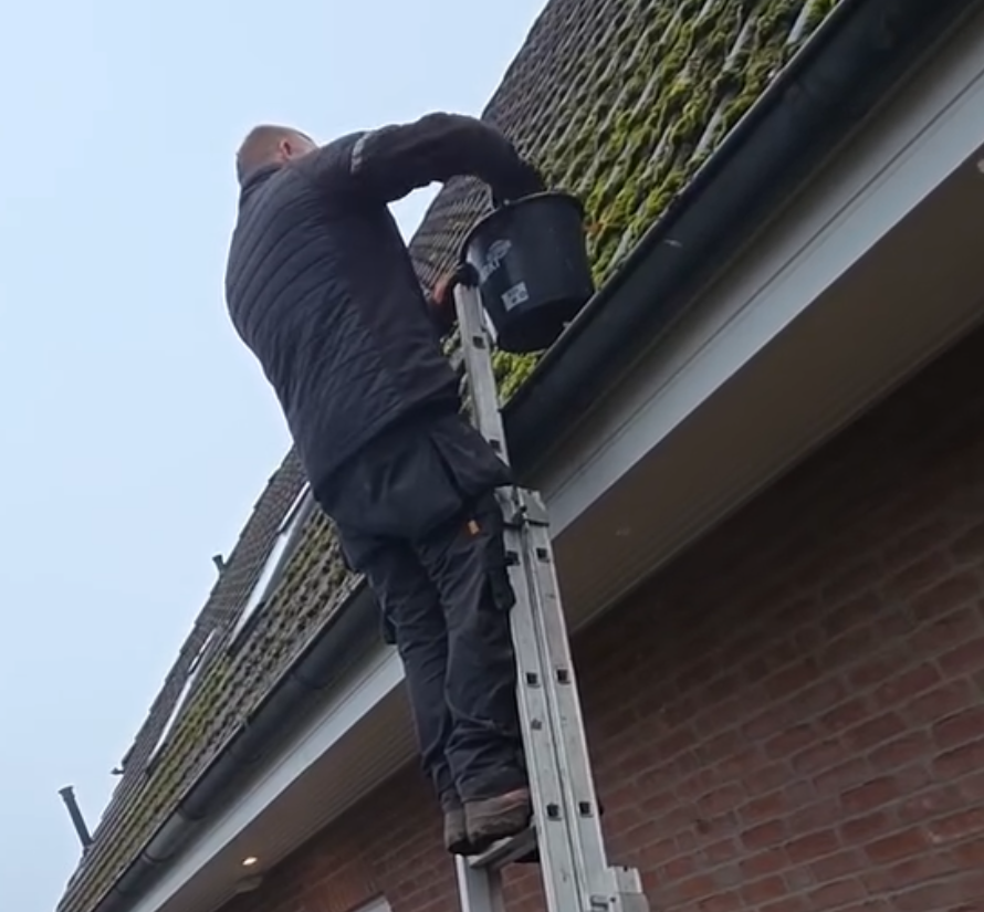 Mann reinigt Dachrinne in Höhe mit Hand und Eimer